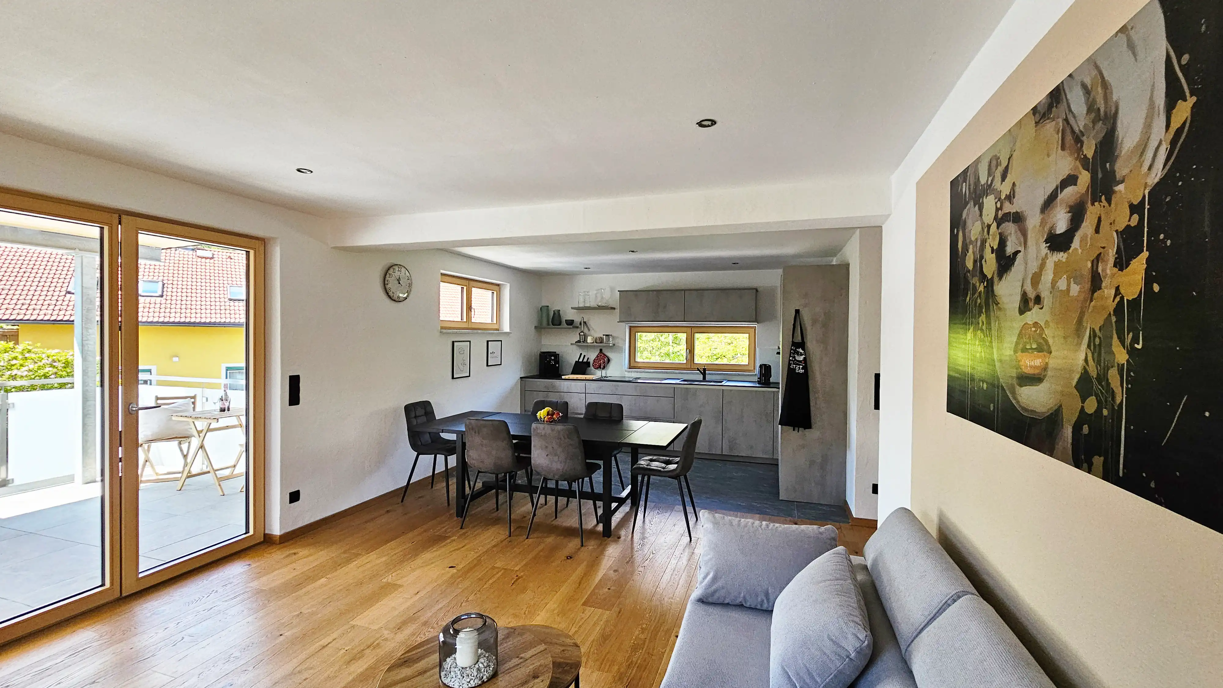 Heller offener Wohn- und Essbereich mit Holzböden, einem grauen Sofa, einem schwarzen Esstisch mit vier Stühlen und einer modernen grauen Küche im Hintergrund. Große Glastüren öffnen sich zu einem Balkon, und ein großes Porträt hängt an der rechten Wand.