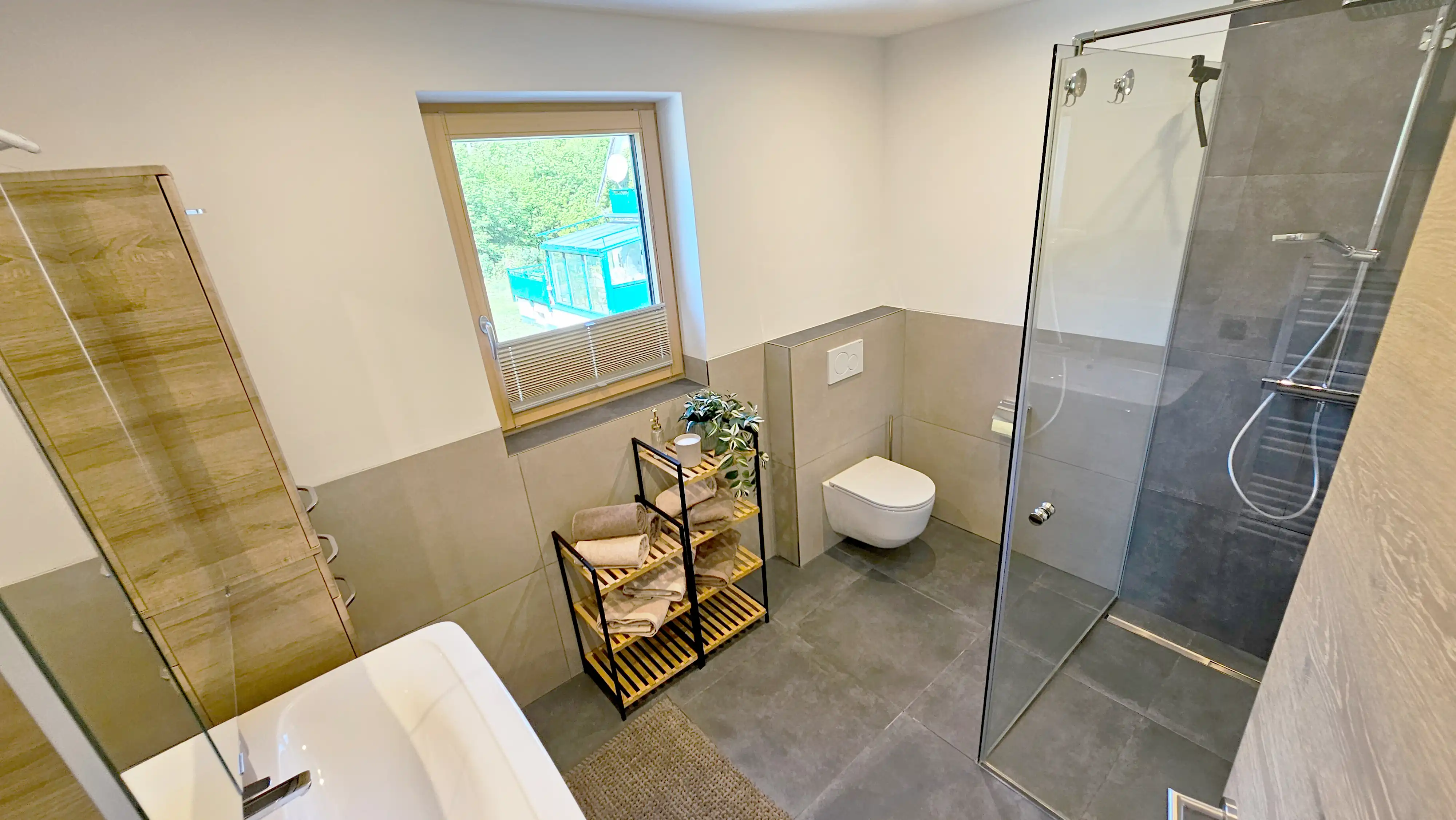 Zeitgenössisches Badezimmer mit grauen Bodenfliesen, einer begehbaren Glasdusche, wandmontierter Toilette, einem Holzregal mit aufgerollten Handtüchern und einem Fenster, das Tageslicht hereinlässt. Neutrale Töne und minimalistische Armaturen schaffen ein sauberes Design.