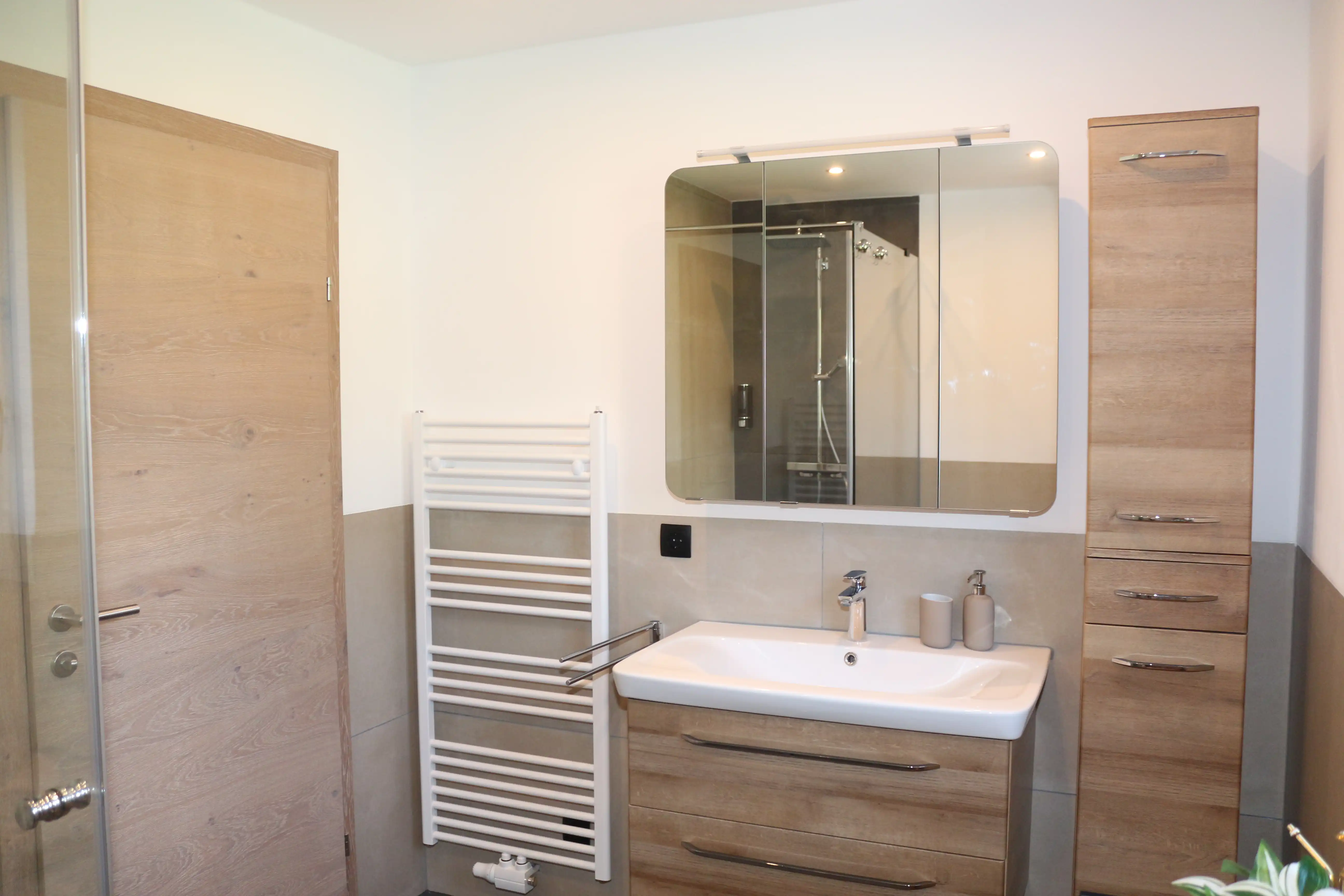 Modernes Badezimmer mit Holzschränken, einem weißen Waschbecken und einem spiegelnden Medizinschrank. Rechts befindet sich ein schwarzes Metall- und Holzregal, das ordentlich aufgerollte braune Handtücher und eine grüne Topfpflanze hält. Ein gewebter Teppich liegt auf dem dunklen Fliesenboden, und Licht strömt durch ein Fenster mit Plisseerollo.