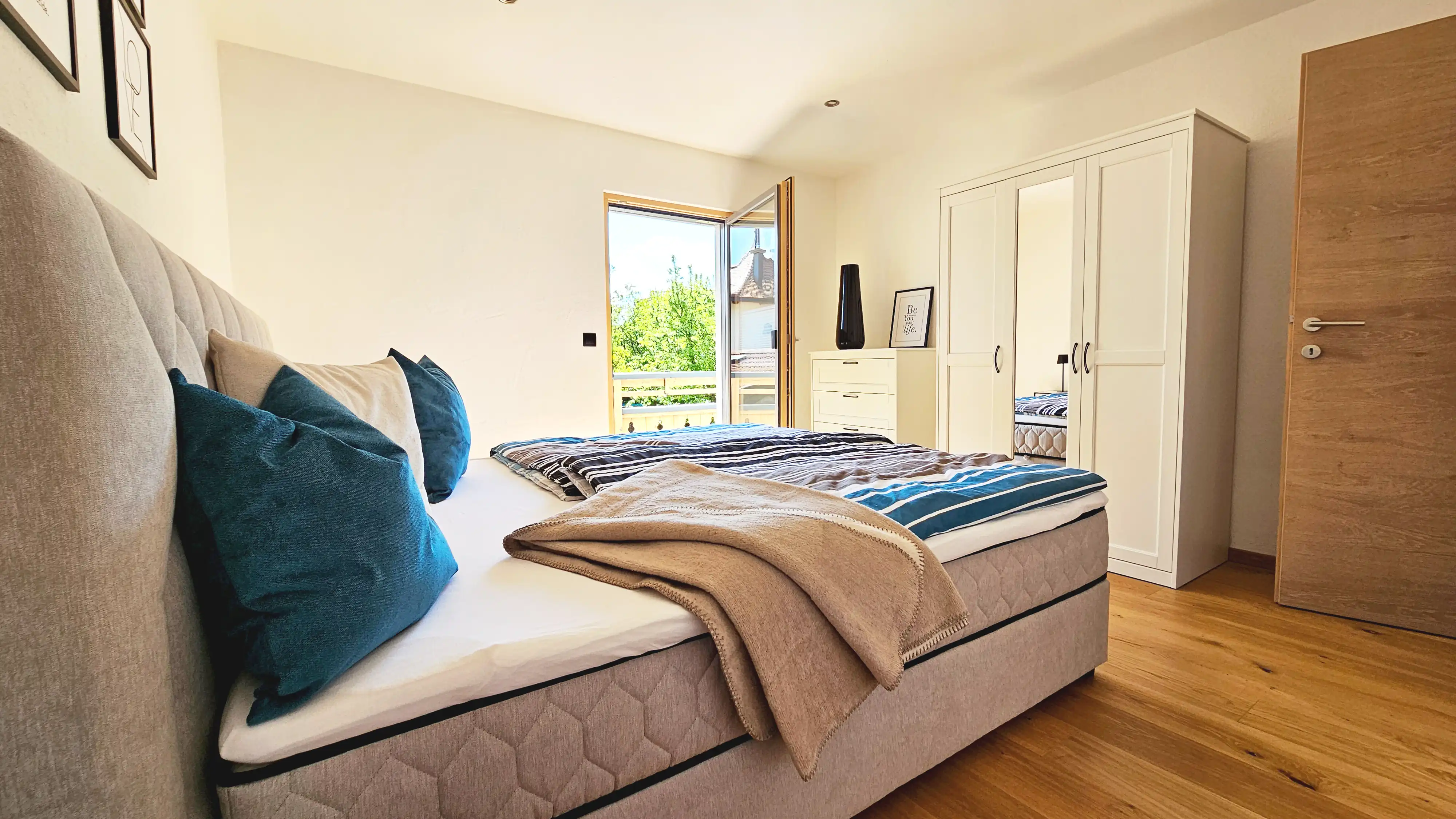 Detailaufnahme des sonnigen Schlafzimmers mit einem beigefarbenen, gepolsterten Boxspringbett, türkis- und beigefarbener gestreifter Bettwäsche und mehreren Zierkissen, gerahmten Bildern an der Wand und einer gläsernen Balkontür.