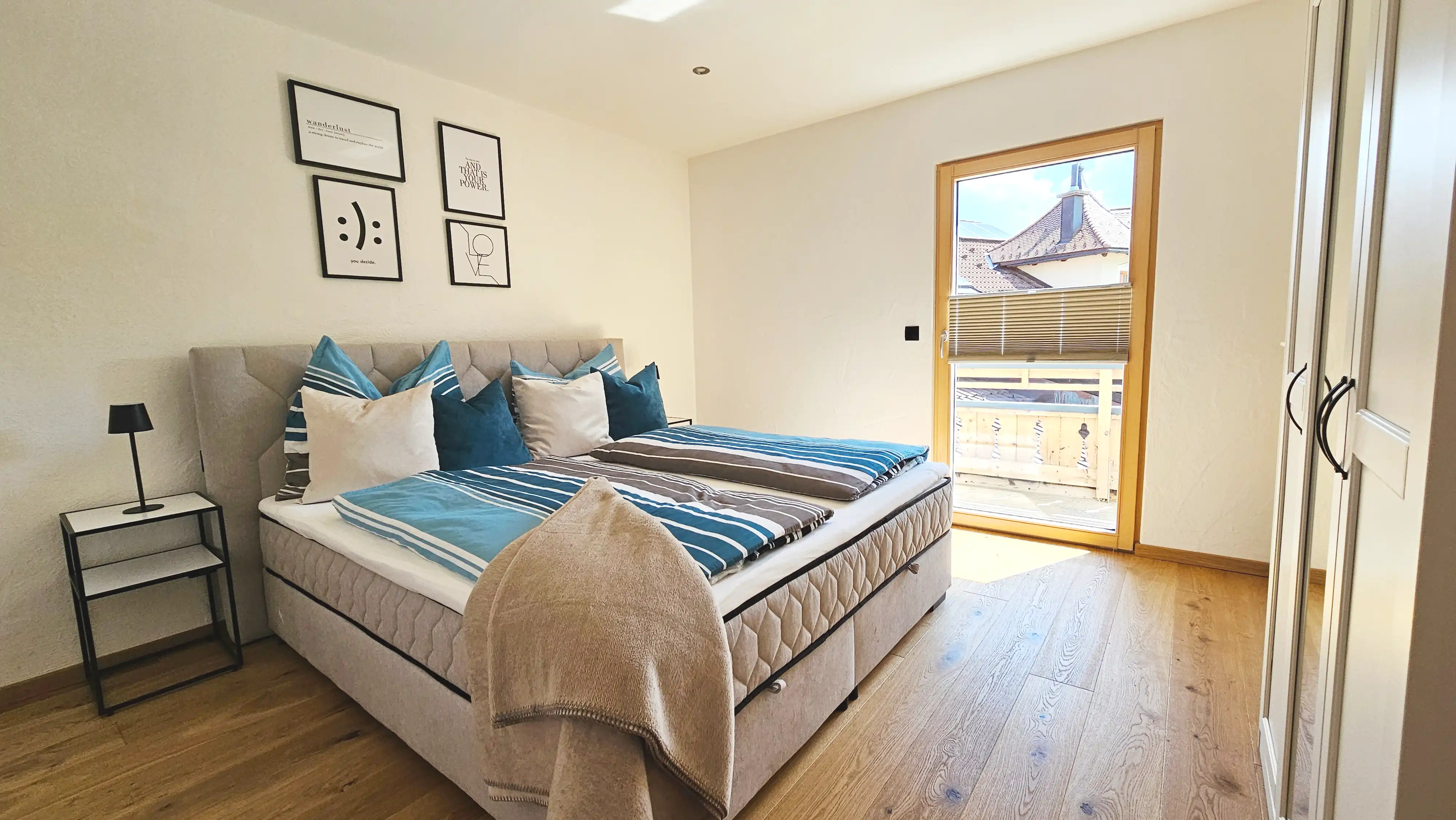 Sonniges Schlafzimmer mit einem beigefarbenen, gepolsterten Boxspringbett mit türkis- und beigefarbener gestreifter Bettwäsche und mehreren Zierkissen, schwarzem Metall-Nachttisch, gerahmten Bildern an der Wand, einer gläsernen Balkontür sowie einem Kleiderschrank entlang der rechten Wand auf Eichenholzboden.