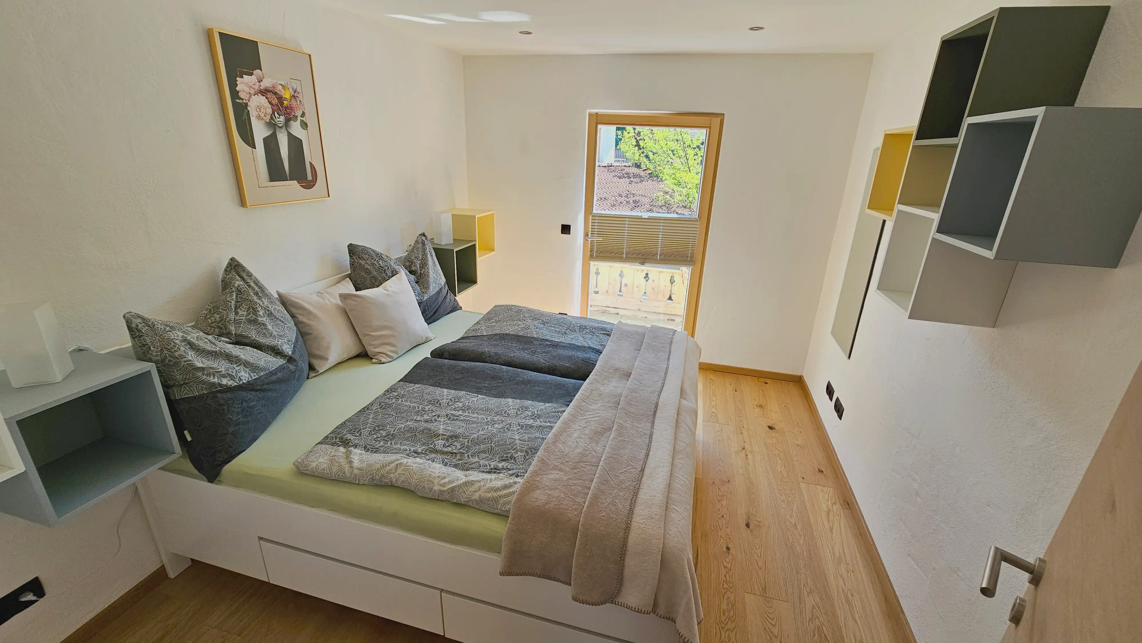 Kleines, helles Schlafzimmer mit einem weißen Plattformbett, das mit grauen, gemusterten Bettdecken und pastellfarbenen Kissen ausgestattet ist, geometrischen Wandregalen, einem gerahmten Blumenbild über dem Bett, einer Glastür zu einem kleinen Balkon und hellen Eichenholzböden.